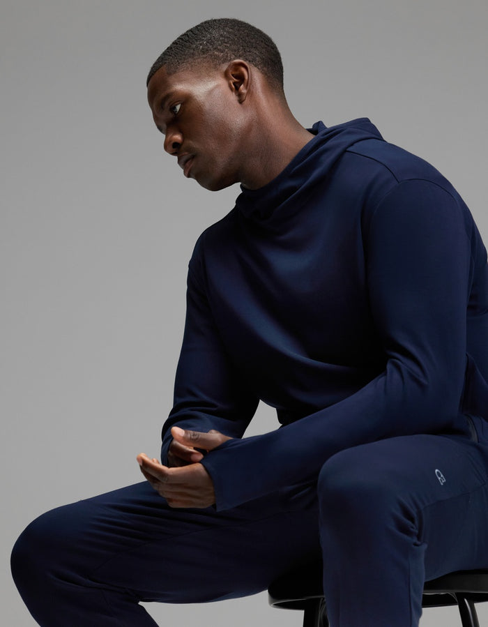 A man wearing Cozy Earth’s Men’s Recovery Jogger in dark blue sits on a stool with his hands clasped, looking down thoughtfully against a plain gray background.|Color:Navy