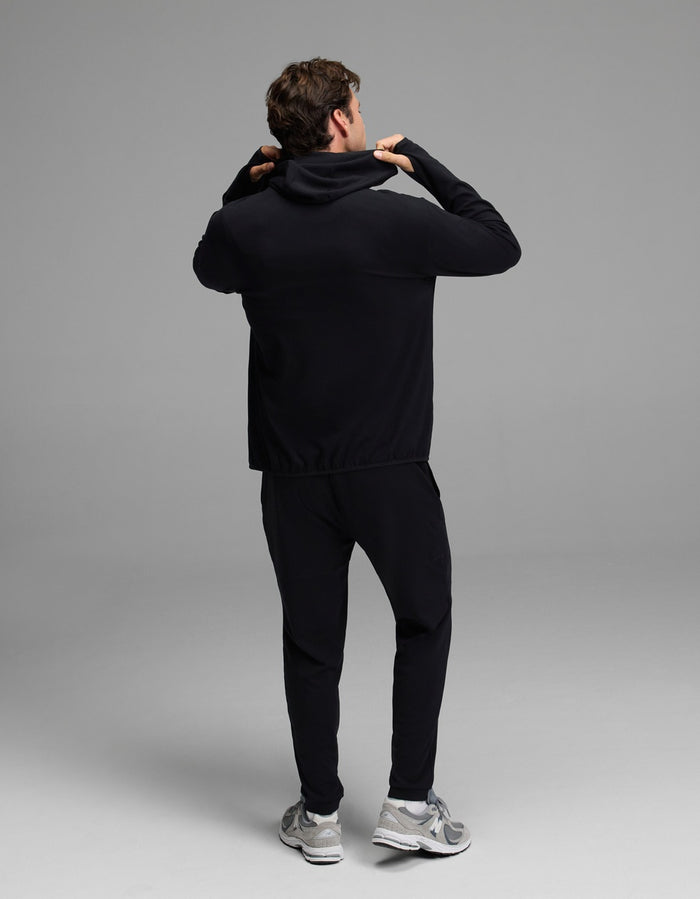 A person faces away while adjusting the hood of a black Men’s Recovery Hoodie & Jogger Set from Cozy Earth, paired with gray sneakers, posing in a simple, well-lit studio against a plain gray background.|Color:Black