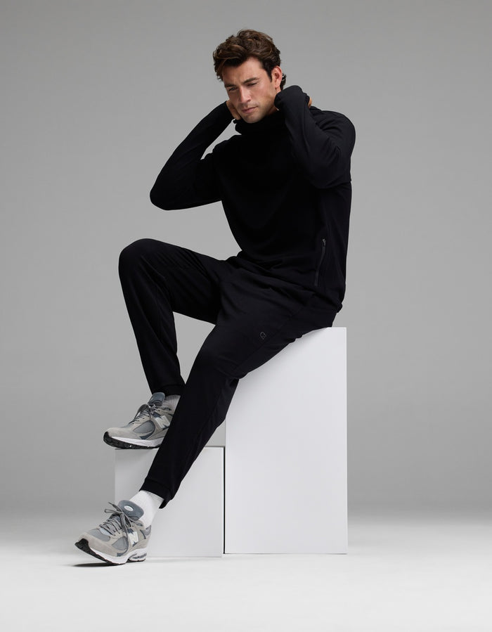 A man wearing Cozy Earth’s Men’s Recovery Jogger and gray sneakers sits on white blocks with one knee raised, hands behind his neck, looking downward against a plain gray background.|Color:Black