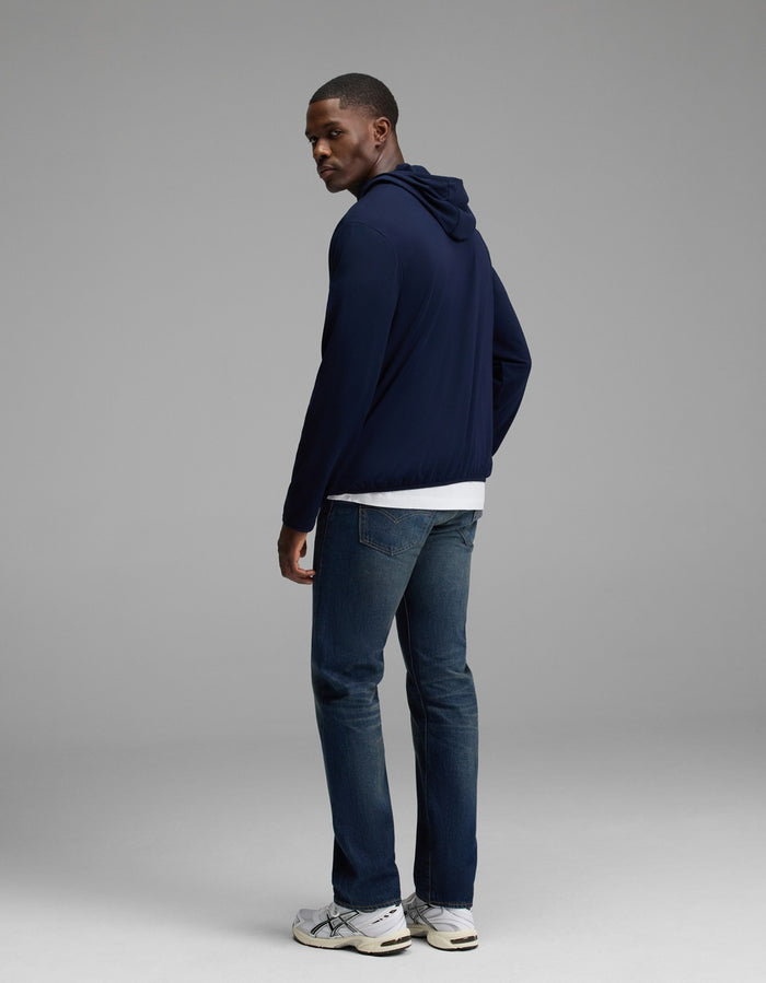 A man stands in a studio, facing slightly away, wearing the Cozy Earth Men’s Recovery Hoodie in navy over a white shirt, paired with blue jeans and white sneakers against a plain gray background.|Color:Navy