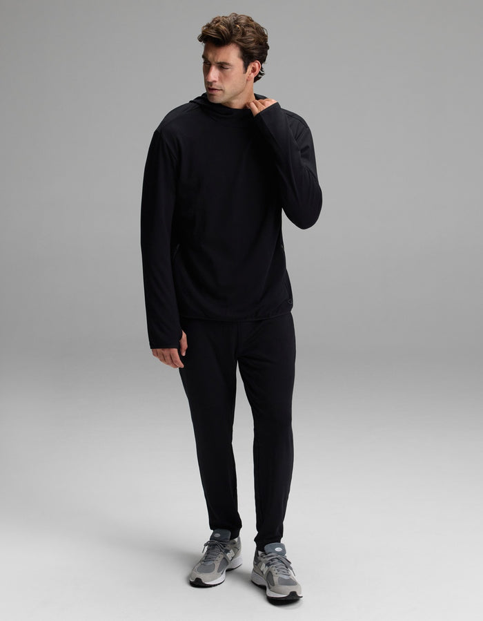 A man wearing the Cozy Earth Men’s Recovery Hoodie in black and matching black pants stands on a gray background, looking to his left with his right hand touching his neck. He completes the look with gray sneakers.|Color:Black