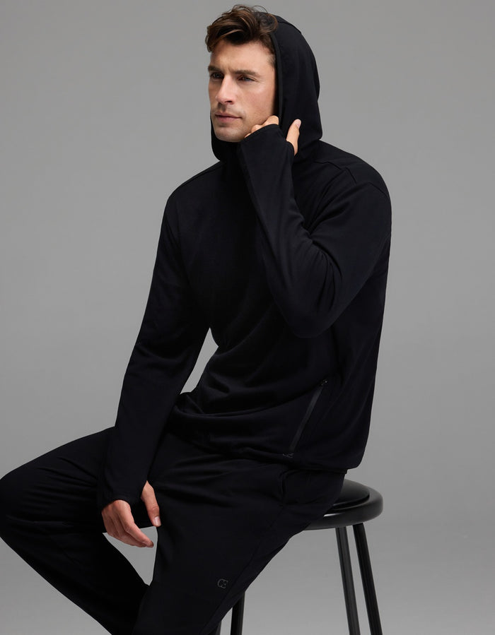 A man wearing the Cozy Earth Men’s Recovery Hoodie and matching pants sits on a black stool against a gray background, with one hand touching his hoodie near his chin.|Color:Black