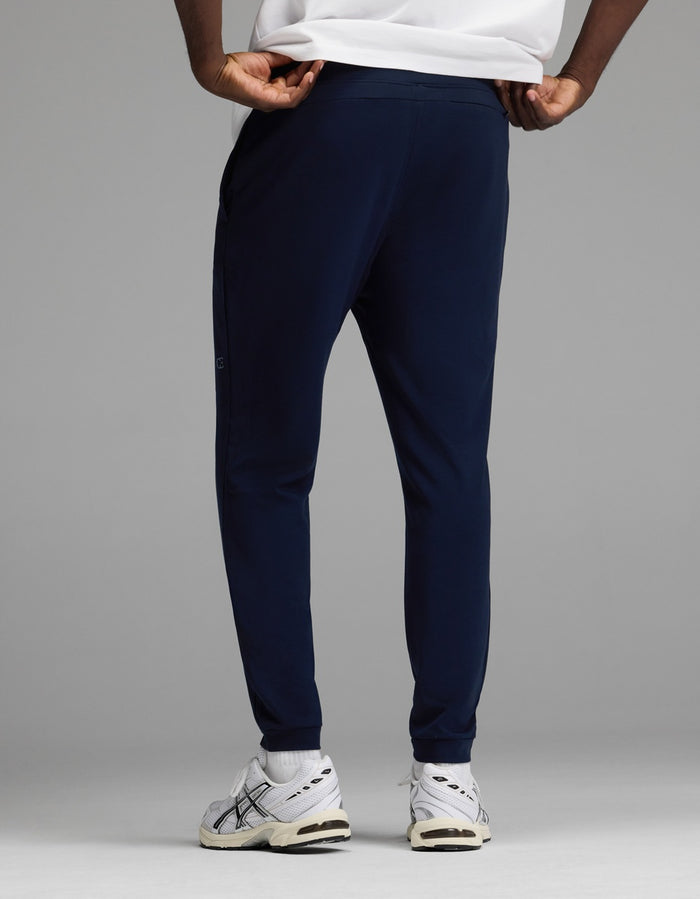 A man models the Cozy Earth Men’s Recovery Jogger in navy, paired with white sneakers featuring black accents and a white shirt, standing against a plain gray backdrop. The photo highlights the back view of the lower body.|Color:Navy