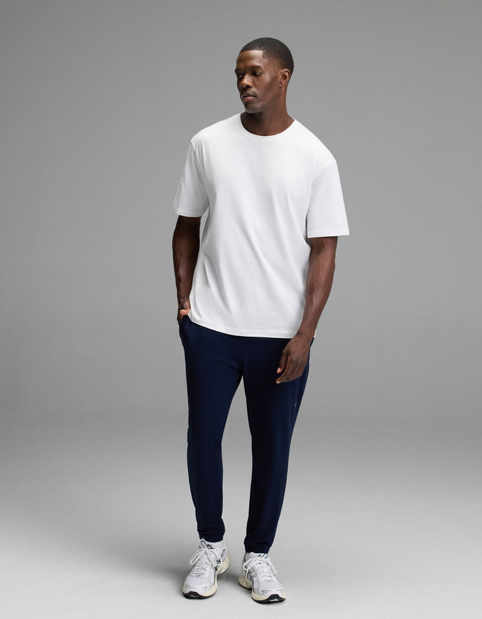 A man stands in a studio wearing a Cozy Earth Men’s Recovery Jogger in dark navy, paired with a plain white t-shirt and white sneakers. He looks slightly to the side with a neutral expression against a plain gray background.|Color:Navy