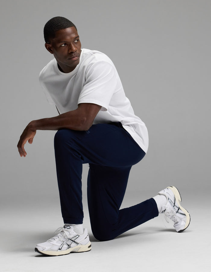 A man wearing a white t-shirt, white sneakers, and Cozy Earth Men’s Recovery Jogger in navy kneels on one knee against a plain gray background, looking to his left.|Color:Navy
