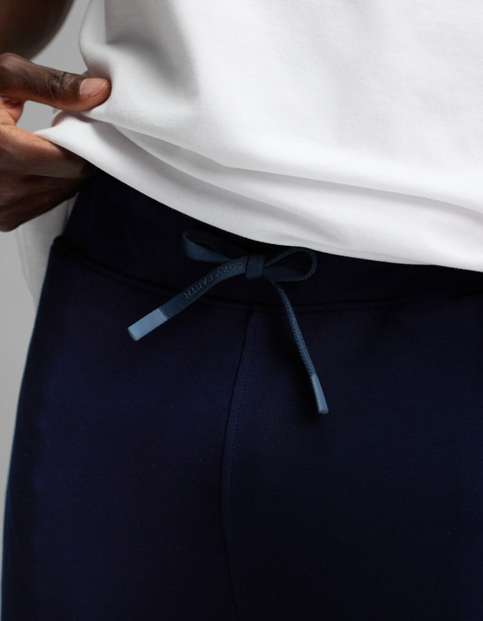 A man wearing Cozy Earth’s Men’s Recovery Jogger in navy blue and a white shirt, adjusting the drawstring waistband with one hand.|Color:Navy