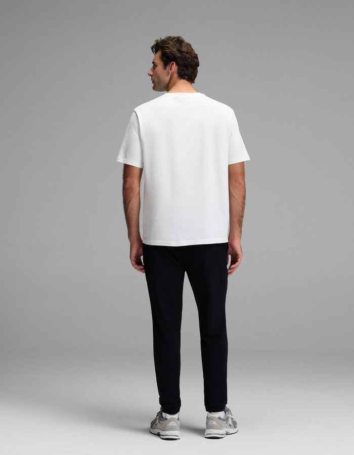 A man stands facing away from the camera, wearing the Cozy Earth Men’s Recovery Jogger with a plain white T-shirt and gray sneakers against a plain gray background.|Color:Black