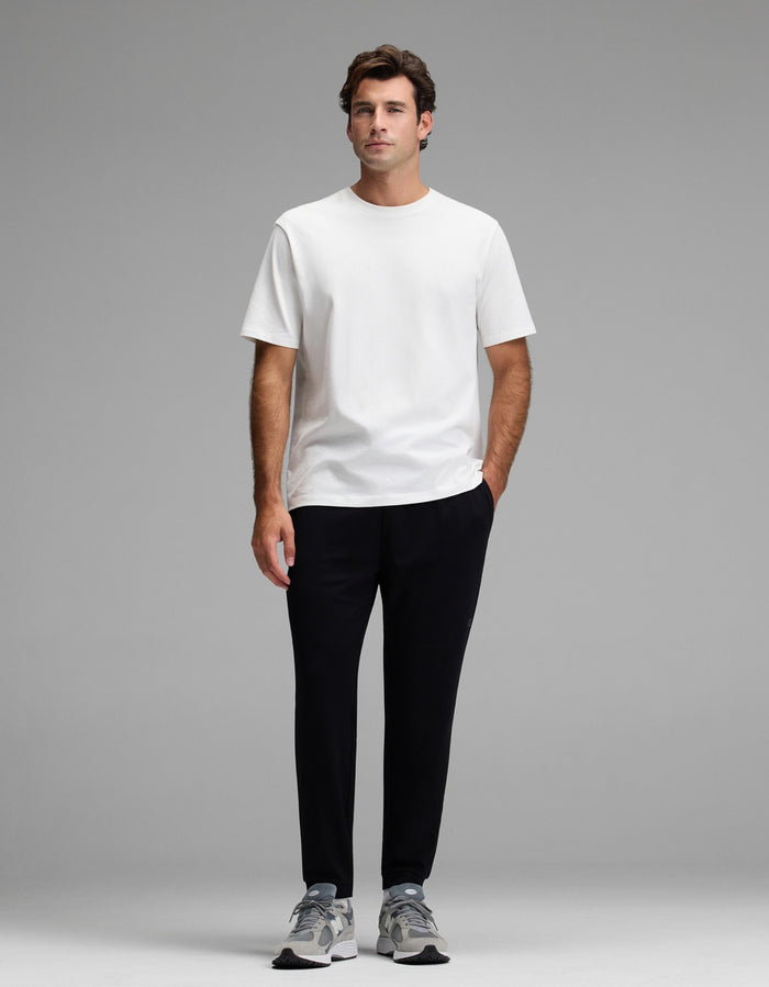 A man stands facing forward against a plain gray background, wearing the Cozy Earth Men’s Recovery Jogger, a plain white t-shirt, and gray sneakers with his hands in his pockets.|Color:Black