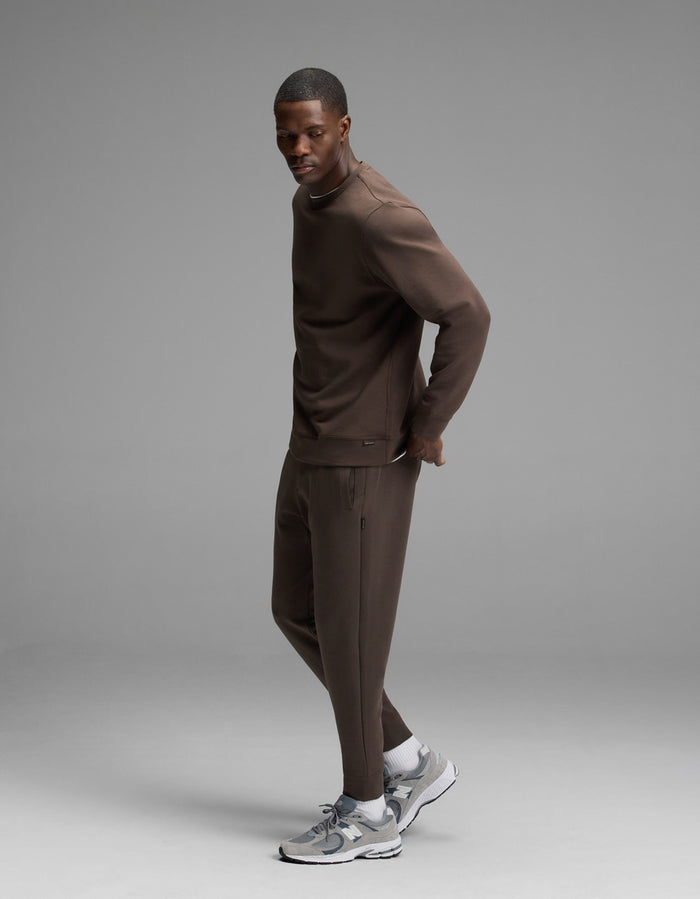 A man wearing the Cozy Earth Men’s StretchTech Crewneck & Jogger Set in brown stands sideways on a gray background, looking toward the camera. He completes his look with gray and white sneakers.|Color:Cacao