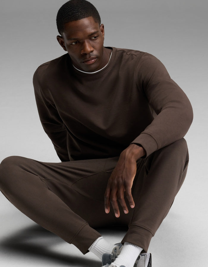 A man sits on the ground in a relaxed pose, wearing the Cozy Earth Men’s StretchTech Crewneck & Jogger Set in brown, paired with white socks and gray sneakers, against a plain gray background.|Color:Cacao