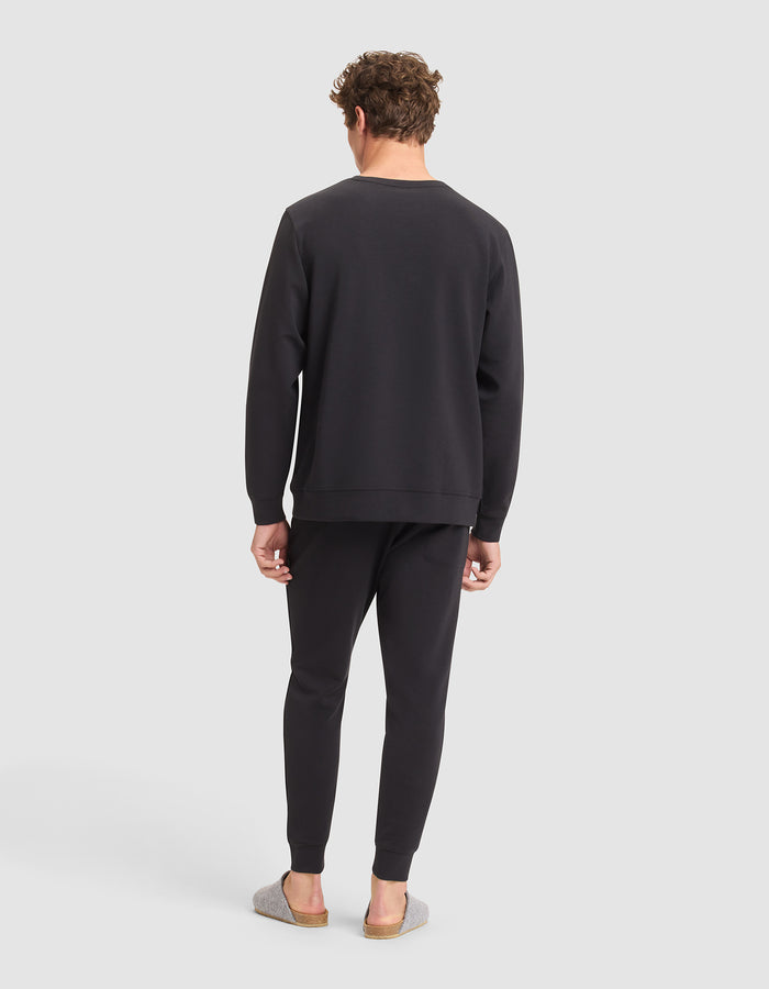 A man faces away from the camera, wearing the Cozy Earth Men's StretchTech Crewneck & Jogger Set in black with gray slip-on shoes, set against a plain light gray background. |Color:Jet Black