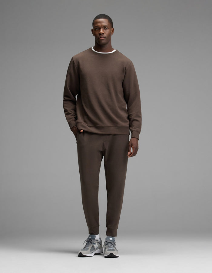 A man stands facing forward, wearing the Cozy Earth Men’s StretchTech Crewneck sweatshirt and matching jogger pants with gray sneakers, against a plain gray background.|Color:Cacao