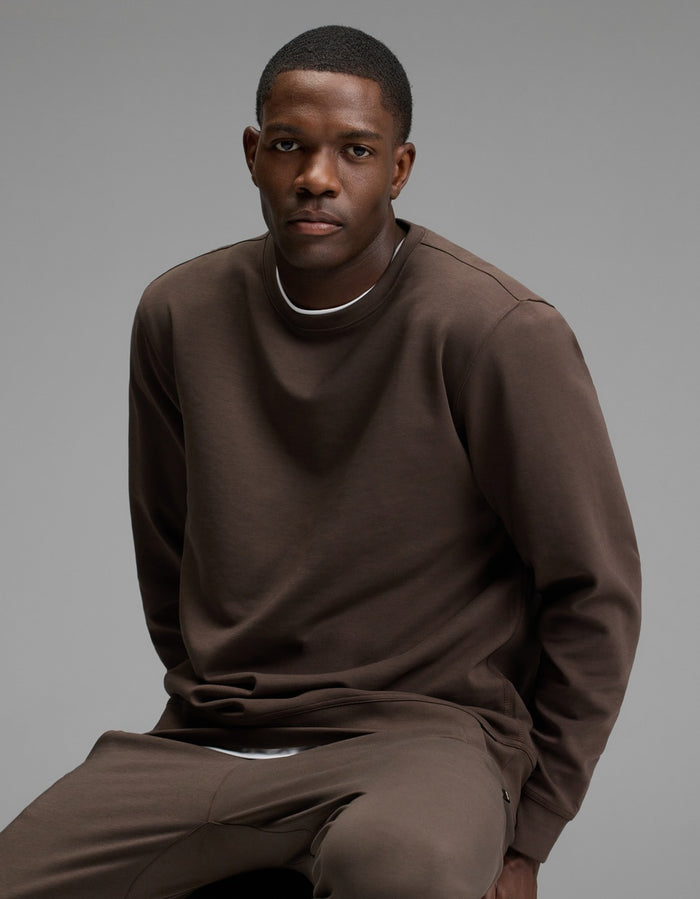 A man sits on a stool wearing the Cozy Earth Men’s StretchTech Crewneck and matching pants, looking at the camera with a neutral expression against a plain gray background.|Color:Cacao