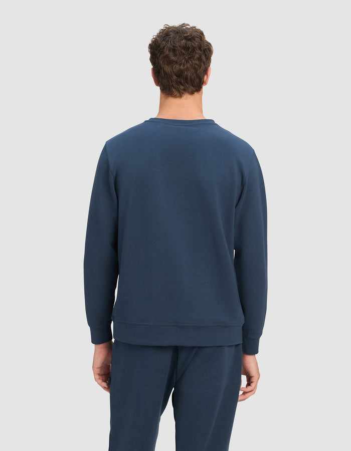 A person with short curly brown hair faces away from the camera, wearing the Cozy Earth Men's StretchTech Crewneck in dark blue against a plain light gray background.|Color:Eclipse