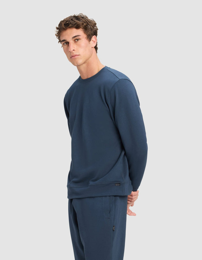 A young man with short, curly brown hair wears a Cozy Earth Men's StretchTech Crewneck in dark blue. He stands against a plain light grey background, looking slightly to the side with his hands behind his back.|Color:Eclipse