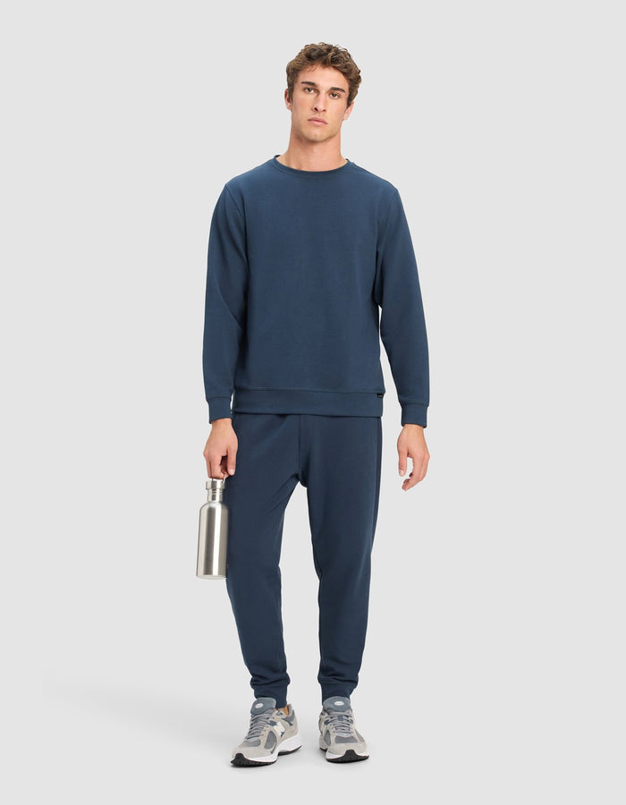 A man faces forward against a light grey background, wearing the Cozy Earth Men's StretchTech Crewneck and matching joggers in navy blue. He holds a silver reusable water bottle and sports grey and white sneakers.|Color:Eclipse