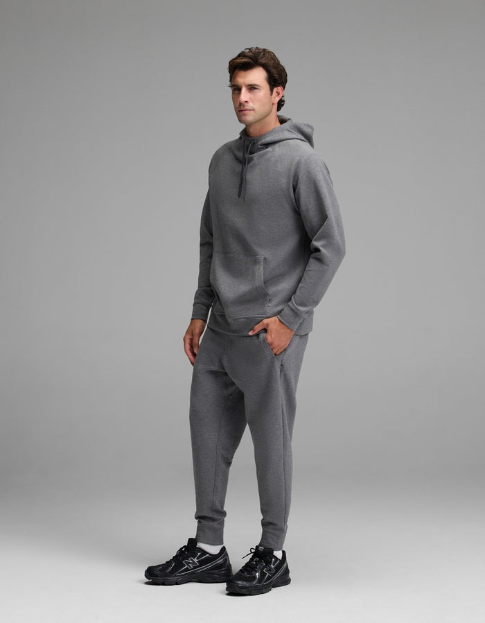A man stands in a studio wearing the Cozy Earth Men’s StretchTech Hoodie & Jogger Set in gray, paired with black sneakers. The background is plain and light gray.|Color:Coal Heather