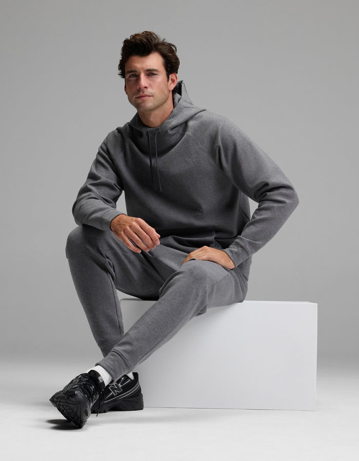 A man models the Cozy Earth Men’s StretchTech Hoodie & Jogger Set in gray, sitting on a white block against a plain gray background. He looks forward with a neutral expression and pairs the outfit with black sneakers.|Color:Coal Heather