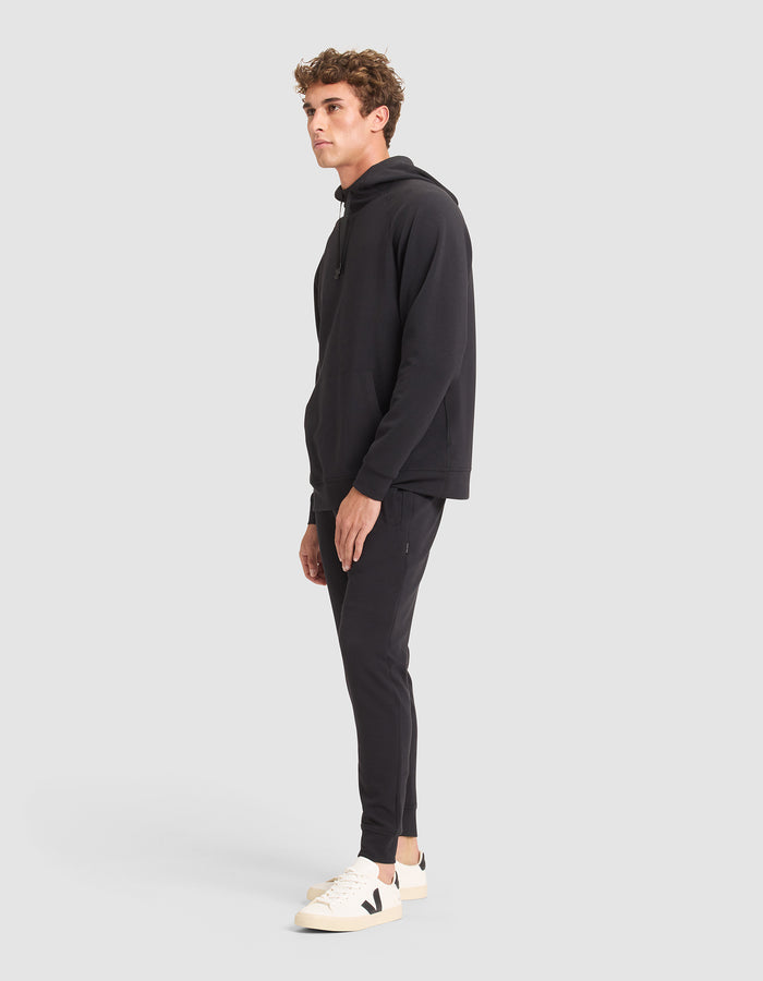 A man stands sideways against a plain light gray background, modeling the Cozy Earth Men's StretchTech Hoodie & Jogger Set in black, paired with white sneakers featuring a black "V" logo. |Color:Jet Black