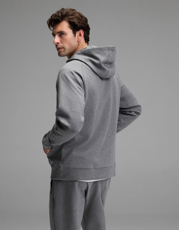A man wearing the Cozy Earth Men’s StretchTech Hoodie and matching sweatpants stands with his back mostly to the camera, turning his head to look over his left shoulder against a plain, light gray background.|Color:Coal Heather