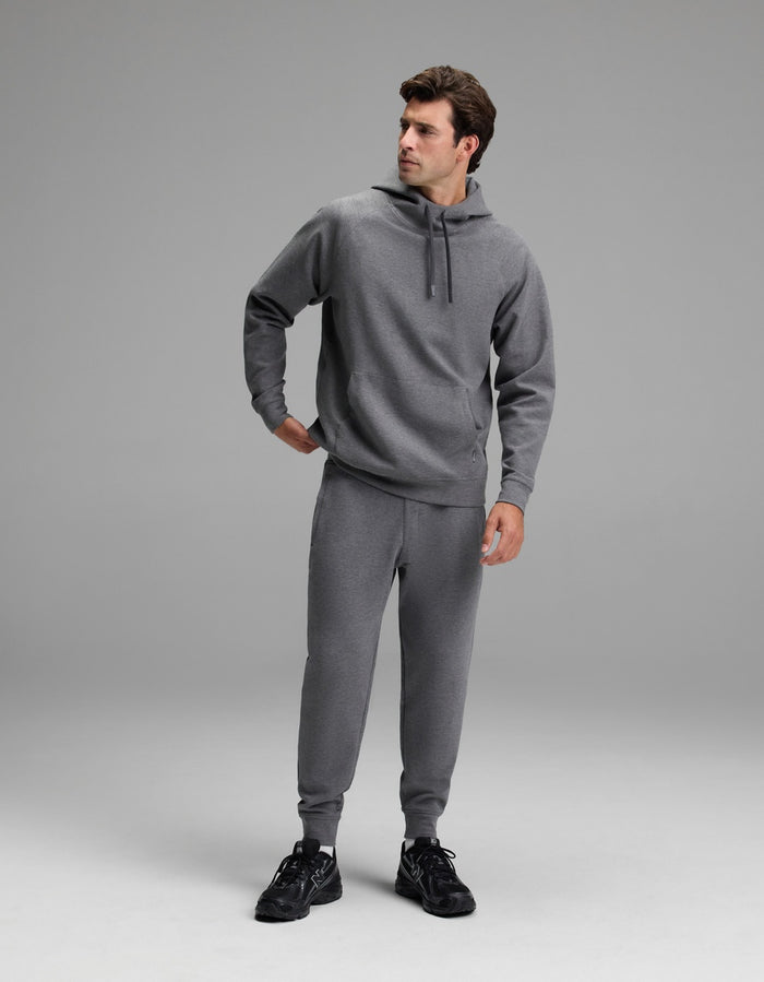 A man stands in a studio wearing the Cozy Earth Men’s StretchTech Hoodie in gray, paired with matching sweatpants and black athletic shoes. He looks to his left with one hand on his hip against a plain, light gray background.|Color:Coal Heather