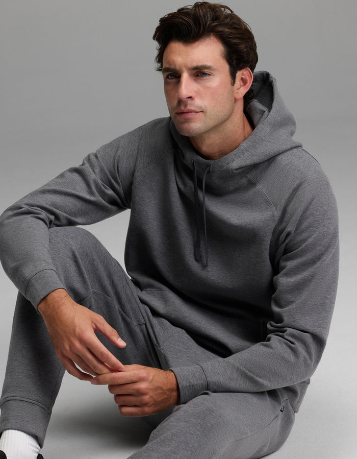 A man wearing the Cozy Earth Men’s StretchTech Hoodie and matching sweatpants sits on the floor against a plain gray background, looking slightly to the side with a neutral expression.|Color:Coal Heather