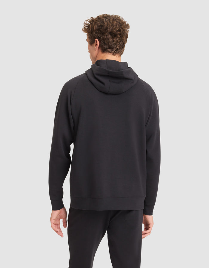 A man with curly hair faces away from the camera, wearing a Cozy Earth Men's StretchTech Hoodie in black and black pants against a light gray background. |Color:Jet Black