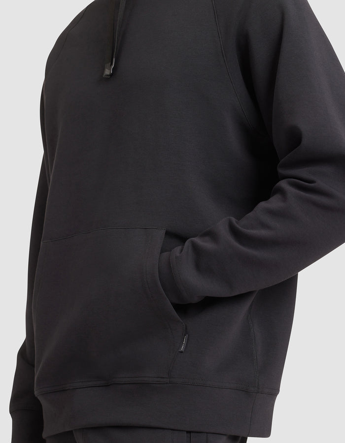 A man models the Cozy Earth Men's StretchTech Hoodie in black, one hand in the front pocket, shown from shoulders to hips against a plain light background. |Color:Jet Black