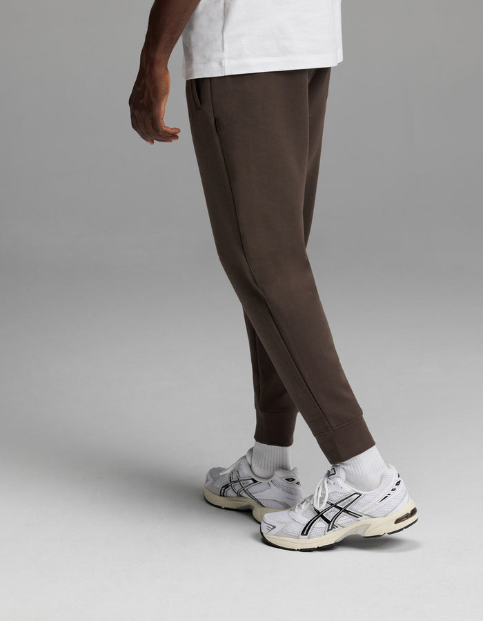 A person is shown from the waist down, standing against a plain gray background, wearing Cozy Earth’s Men’s StretchTech Jogger in brown, paired with a white T-shirt, white socks, and white sneakers.|Color:Cacao