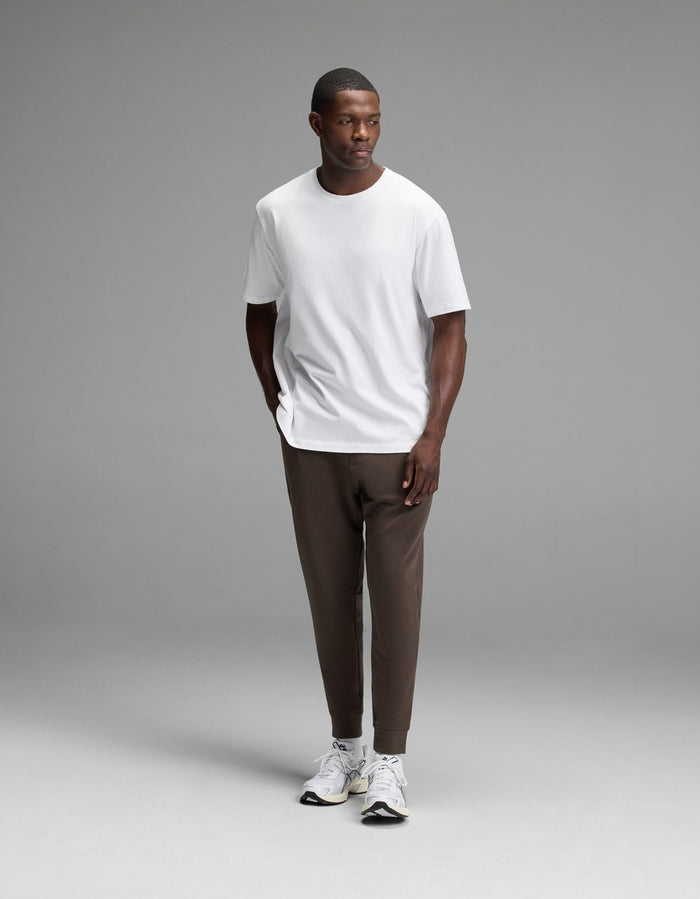 A man stands against a plain gray background wearing a white T-shirt, Cozy Earth Men’s StretchTech Jogger in brown, and white sneakers, with his hands in his pockets.|Color:Cacao