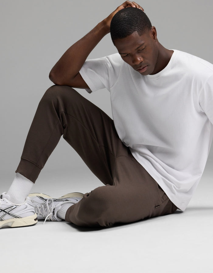 A man sits on the floor wearing a white t-shirt, white socks, gray sneakers, and Cozy Earth’s Men’s StretchTech Jogger in brown. He rests one hand on his head and looks down thoughtfully against a plain background.|Color:Cacao