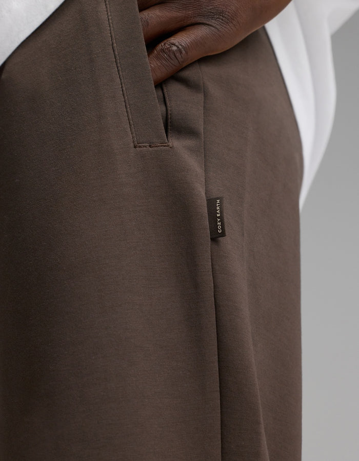 A close-up of a person’s hand in the pocket of Cozy Earth Men’s StretchTech Jogger pants in brown, featuring a small fabric tag with text. The person is also wearing a white shirt.|Color:Cacao