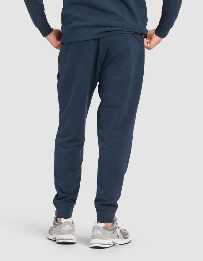 A person shown from the back wearing the Cozy Earth Men's StretchTech Quarter Zip & Jogger Set in navy blue with light gray athletic shoes, standing against a plain light background.|Color:Eclipse