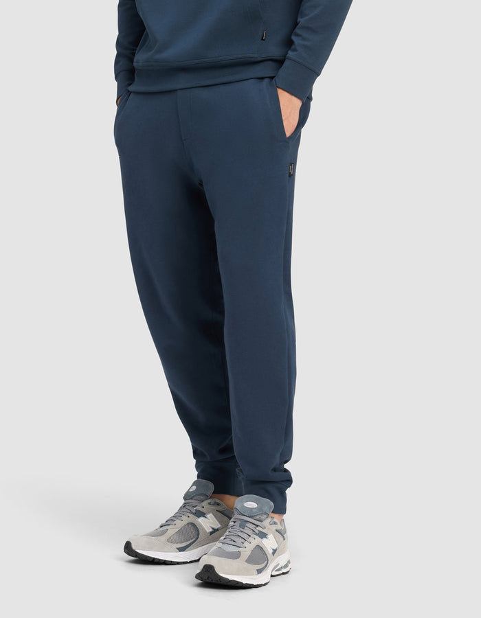 A man wears the Cozy Earth Men's StretchTech Quarter Zip & Jogger Set in navy blue, with hands in pockets, paired with gray and white sneakers, standing against a plain light background.|Color:Eclipse