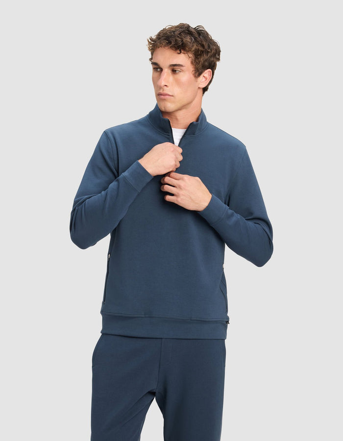 A young man stands against a plain light gray background, adjusting the zipper on his Cozy Earth Men's StretchTech Quarter Zip in dark blue, paired with matching pants.|Color:Eclipse