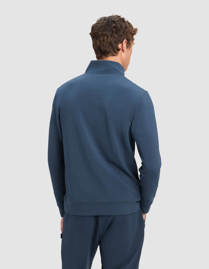 A man with curly brown hair is seen from behind wearing the Cozy Earth Men's StretchTech Quarter Zip in blue with matching pants, standing against a plain light gray background.|Color:Eclipse