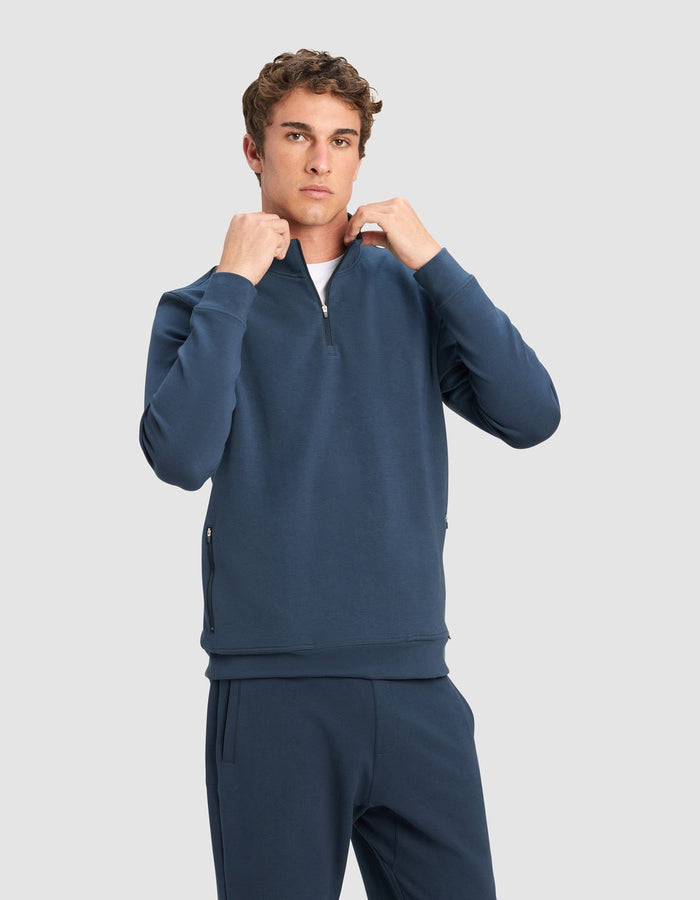 A young man with short curly hair wears the Cozy Earth Men's StretchTech Quarter Zip and matching joggers, standing against a plain light gray background and adjusting the zipper with both hands.|Color:Eclipse