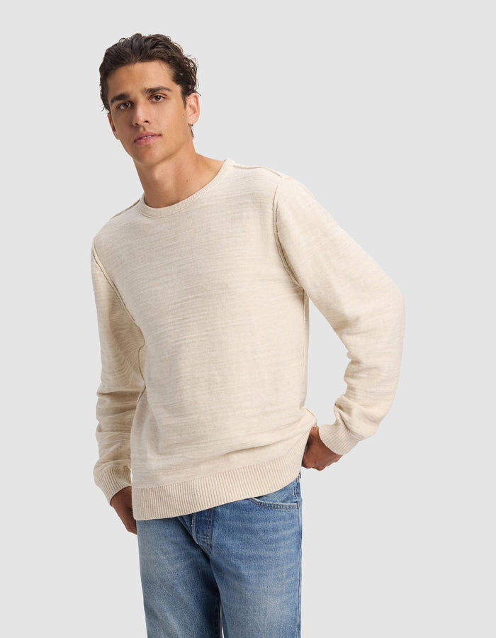 A young man wears the Cozy Earth Men's Tate Crewneck in light beige with blue jeans, standing against a plain gray background with one hand on his hip and a neutral expression.|Color:Alabaster