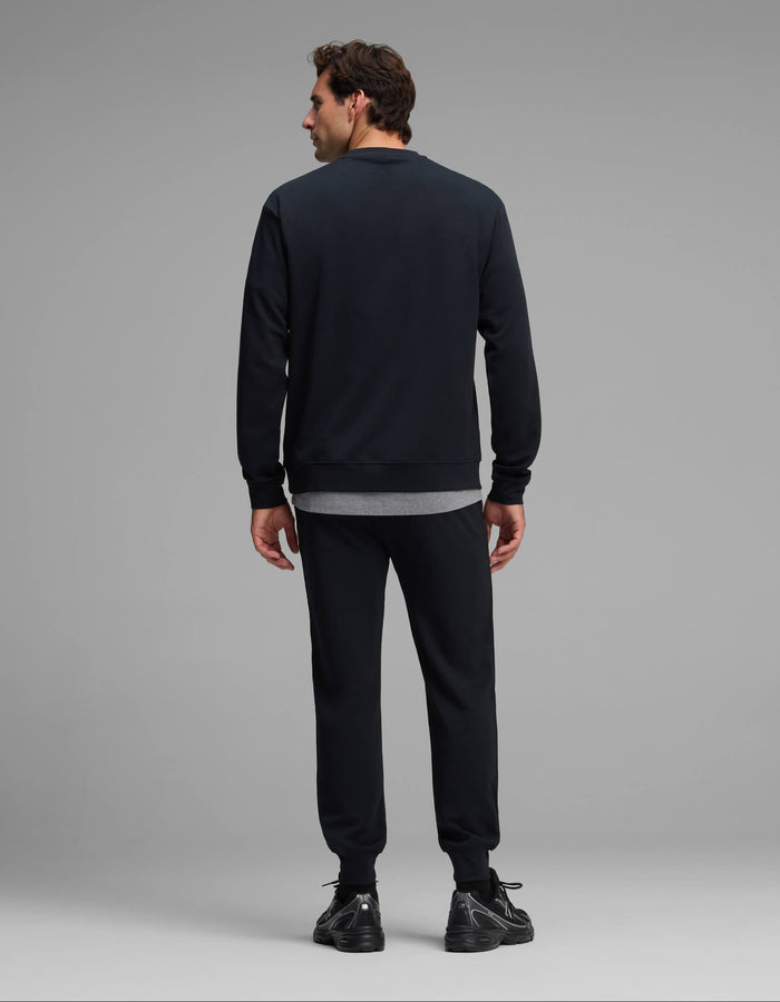 A man is standing with his back to the camera, wearing the Cozy Earth Men's Versa Crewneck & Jogger Set in black, paired with black sneakers and a gray shirt peeking out underneath. The background is plain and light gray.|Color:Jet Black