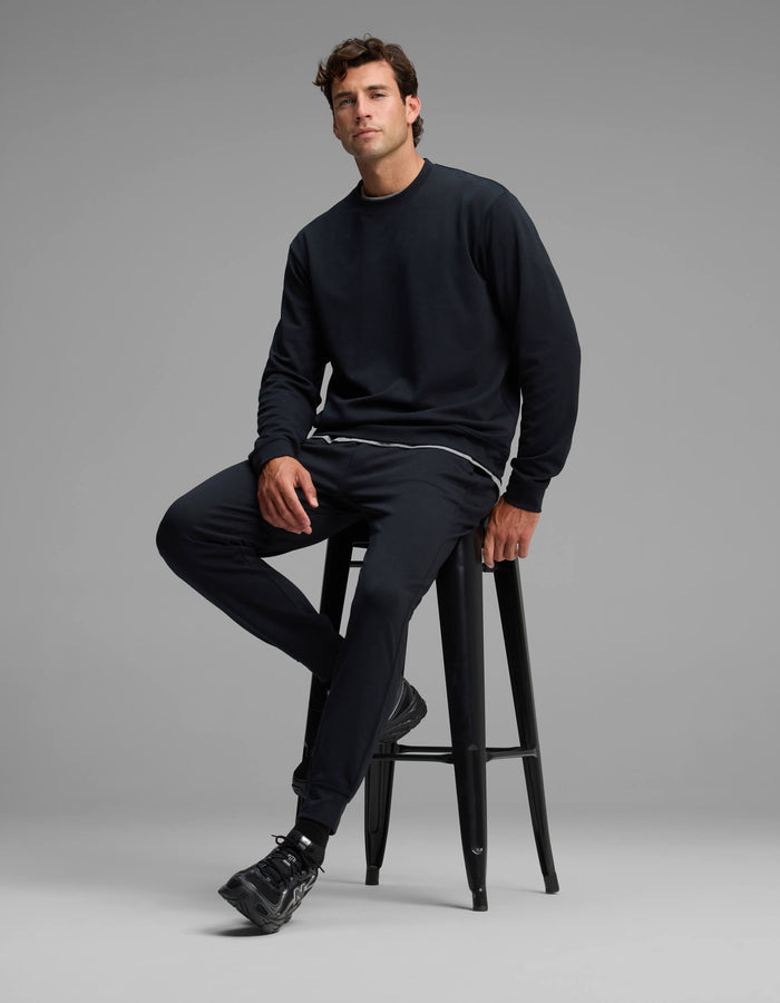 A man dressed in the Cozy Earth Men's Versa Crewneck & Jogger Set sits on a tall black stool against a plain gray background.|Color:Jet Black