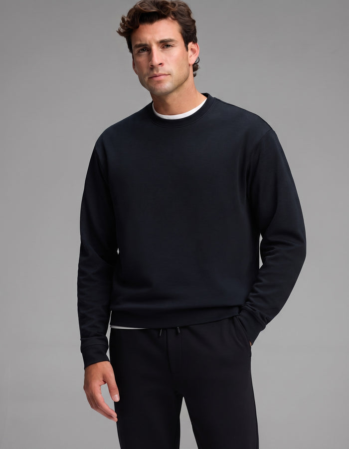 A man with short brown hair wears the Cozy Earth Men's Versa Crewneck & Jogger Set in black over a white shirt, standing against a plain gray background and looking at the camera with a neutral expression.|Color:Jet Black