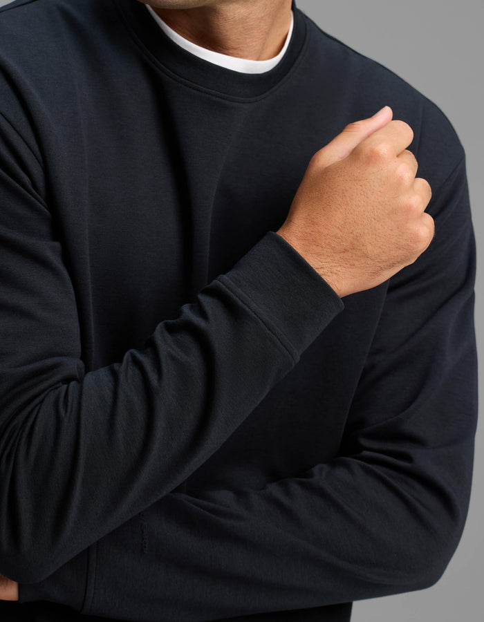 A person is shown from the shoulders down wearing the Cozy Earth Men's Versa Crewneck in black, with one arm crossed and the other bent, hand near the chest, against a gray background.|Color:Jet Black