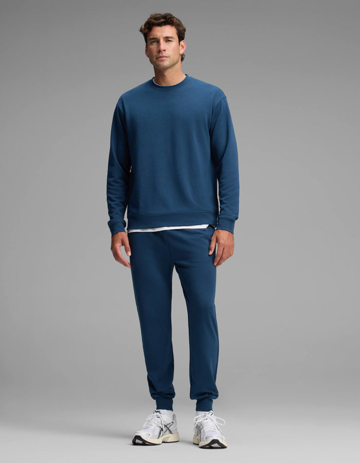 A man stands facing forward against a plain gray background, wearing the Cozy Earth Men's Versa Crewneck & Jogger Set in blue, paired with white sneakers and a white shirt visible beneath the sweatshirt.|Color:Obsidian