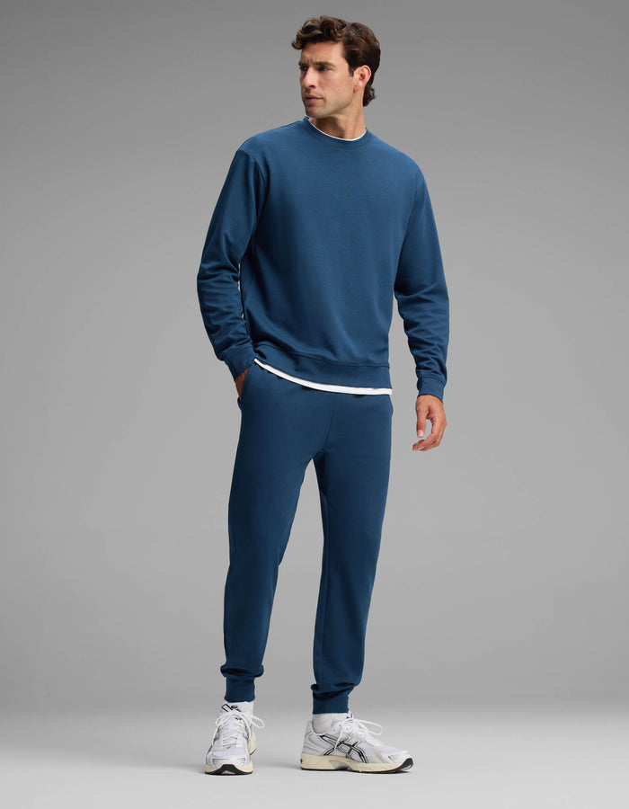 A man wearing the Cozy Earth Men's Versa Crewneck & Jogger Set in blue, paired with a white undershirt and white sneakers, stands against a plain gray background, looking to his right.|Color:Obsidian