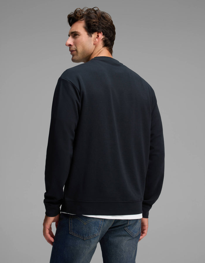 A man with dark hair models the Cozy Earth Men's Versa Crewneck in black with blue jeans, standing with his back to the camera and looking over his left shoulder against a plain gray background.|Color:Jet Black