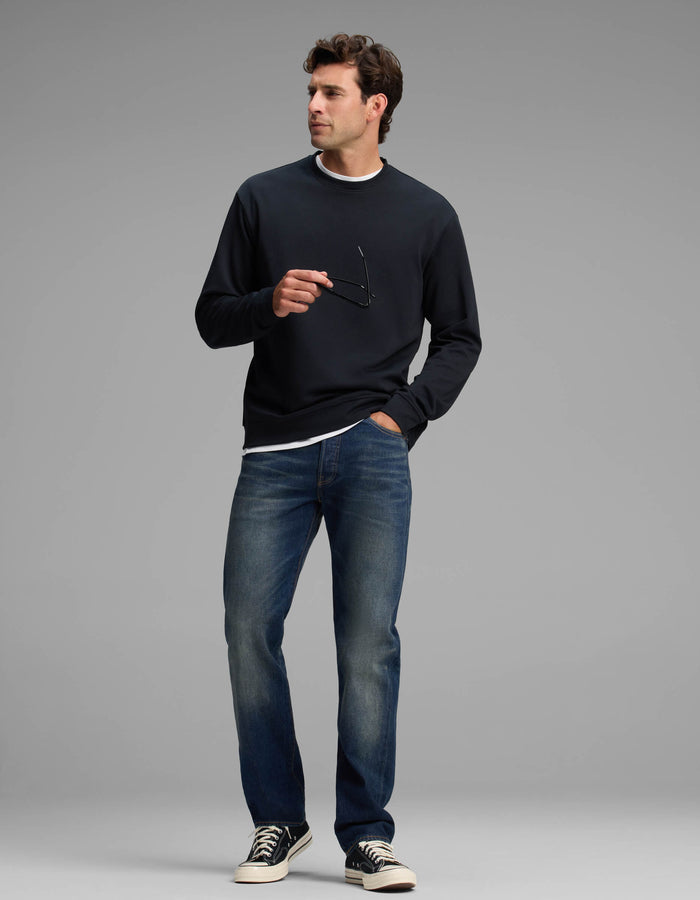 A man wearing the Cozy Earth Men's Versa Crewneck, paired with blue jeans and black sneakers, stands holding eyeglasses in one hand while looking to the side against a plain gray background.|Color:Jet Black