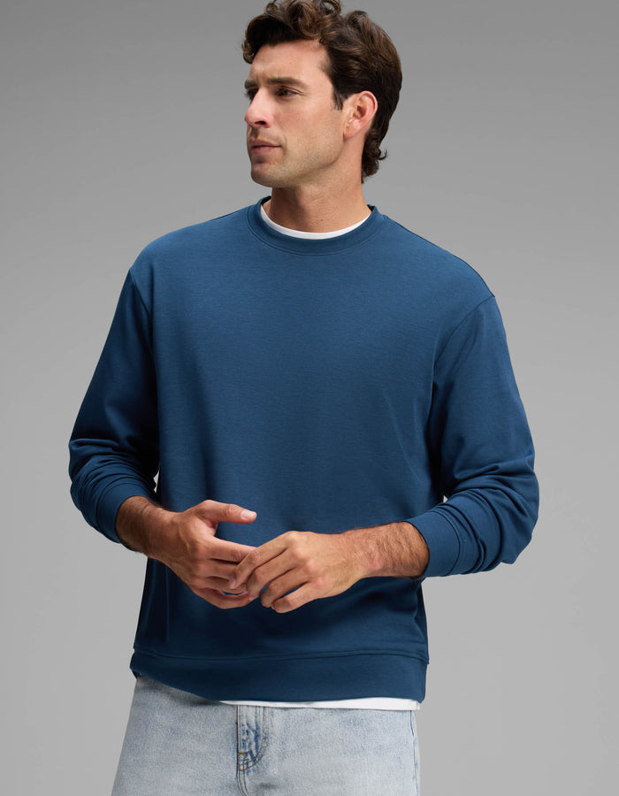 A man wearing the Cozy Earth Men's Versa Crewneck in blue over a white shirt and light blue jeans stands against a plain gray background, looking to his left with his hands loosely clasped.|Color:Obsidian