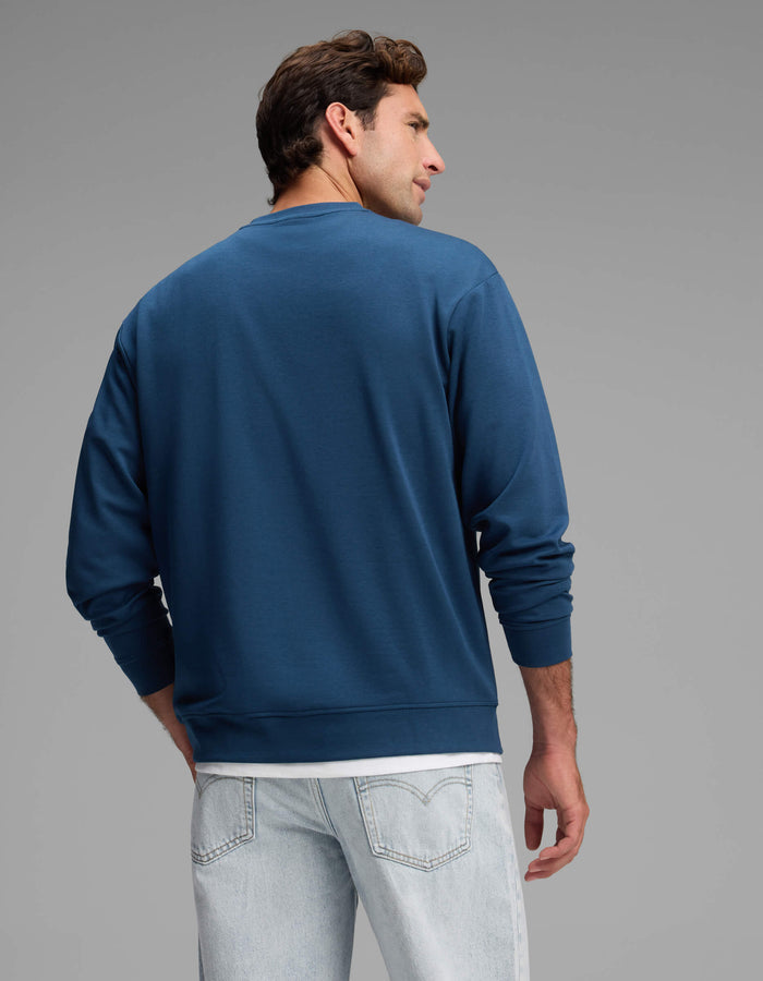 A man is shown from behind, wearing Cozy Earth’s Men's Versa Crewneck in blue over a white t-shirt and light-wash jeans, standing against a plain gray background.|Color:Obsidian