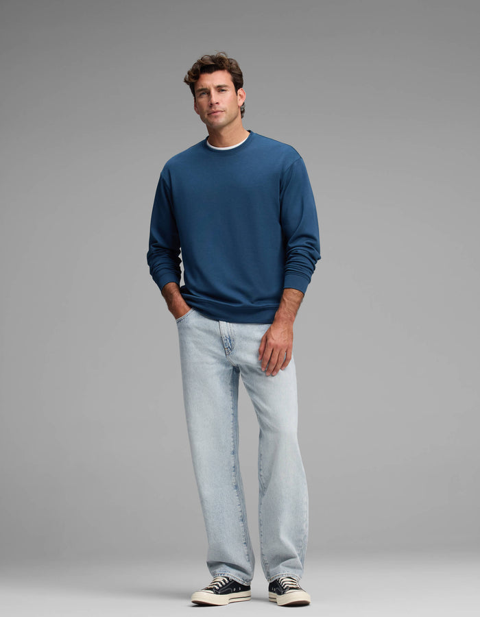 A man stands against a plain gray background wearing the Cozy Earth Men's Versa Crewneck, light blue jeans, and black-and-white sneakers.|Color:Obsidian