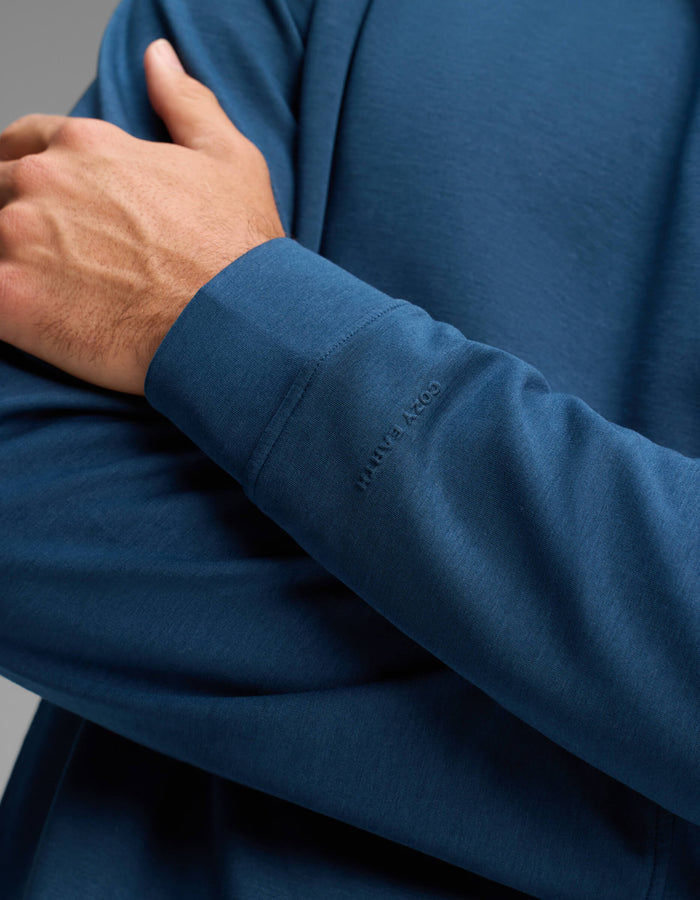 A person wearing the Men's Versa Crewneck by Cozy Earth in blue folds one arm across their chest. The shirt features a subtle "COZY EARTH" embossing near the wrist.|Color:Obsidian
